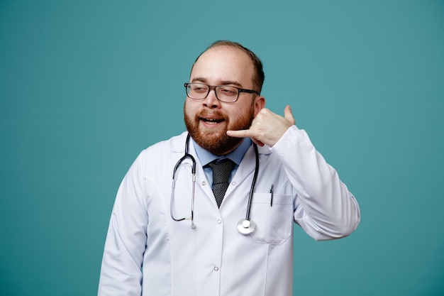 Pleased young male doctor wearing glasses medical coat and stethoscope around his neck looking at side showing call gesture isolated on blue background