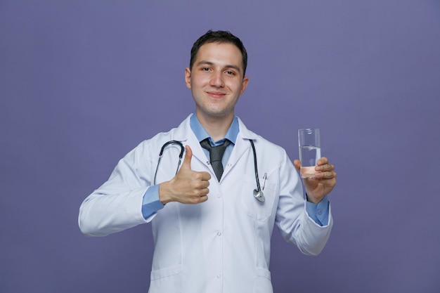 Pleased young male doctor wearing medical robe and stethoscope around neck showing glass of water looking at camera showing thumb up isolated on purple background