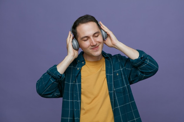 pleased young male student wearing headphones keeping hands on headphones listening to music with closed eyes isolated on purple background