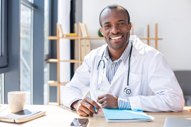 Pleased young man keeping smile on face while sitting at his workplace