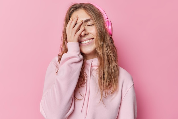 Pleased young woman covers face with hand smiles broady closes eyes has upbeat mood dressed in casual hoodie listens audio track via headphones isolated over pink background. Emotions concept