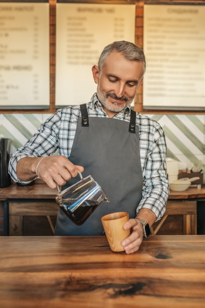 Pleasure. Smiling middle aged man in apron standing at bar counter with pleasure pouring coffee into cup in his cafe