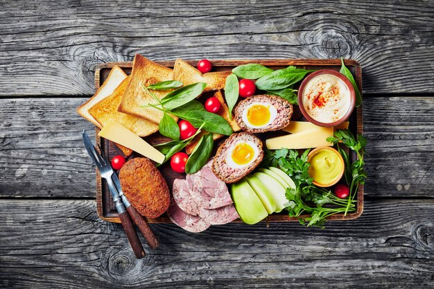 Photo ploughman's lunch of toasts, cheddar cheese wedges, apple, scotch eggs, headcheese, tomatoes, greens, hot mustard served on a wooden tray on a rustic table, view from above, flatlay