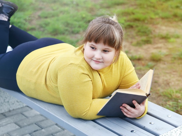 Plump smiling girl lying on bench in park holds book in her hands and looks away