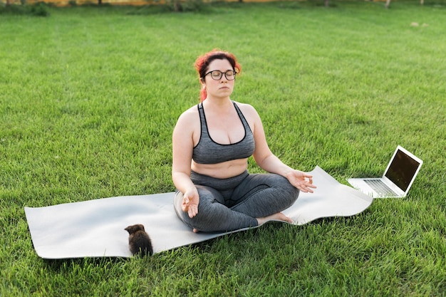 Plus size body positivity woman doing meditation on yoga mat with kitten on backyard wellness and mental health concept