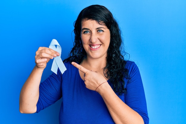 Plus size brunette woman holding blue ribbon smiling happy pointing with hand and finger