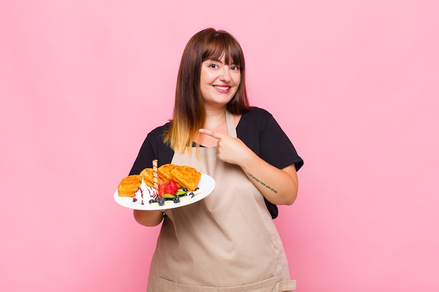 Plus size woman smiling cheerfully, feeling happy and pointing to the side and upwards, showing object in copy space