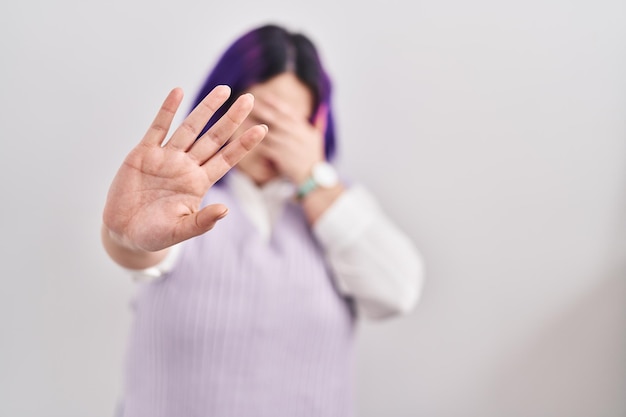 Plus size woman wit purple hair standing over white background covering eyes with hands and doing stop gesture with sad and fear expression. embarrassed and negative concept.