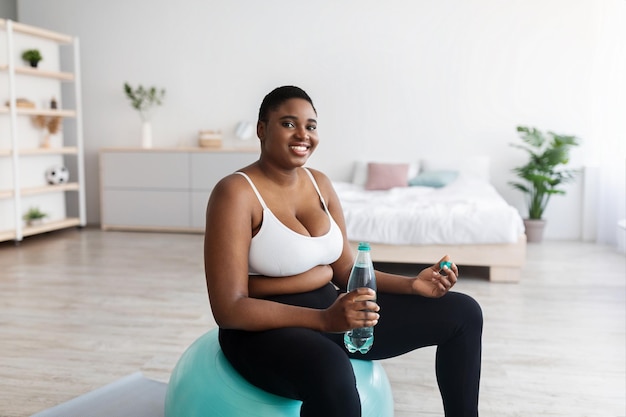 Plus size young black woman sitting in fitness ball with bottle of water exercising at home during