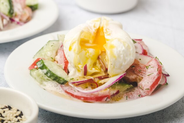 Poached egg on chopped and mixed vegetables with sour cream, white dishes, gray, stone background, close up