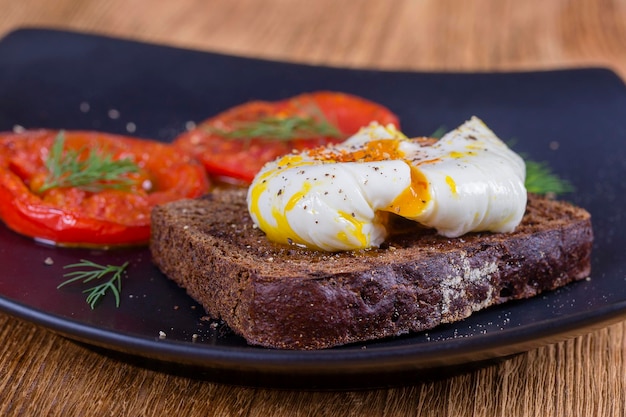 Poached egg on a piece of bread with fried red tomatoes on a wooden table close up