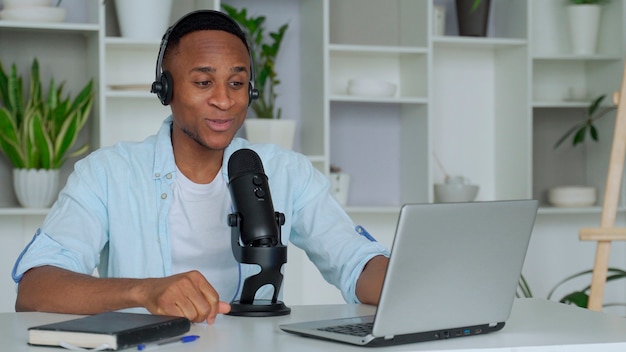 Podcast concept  happy young black man audio blogger in headphones with laptop computer and microphone broadcasting at home off