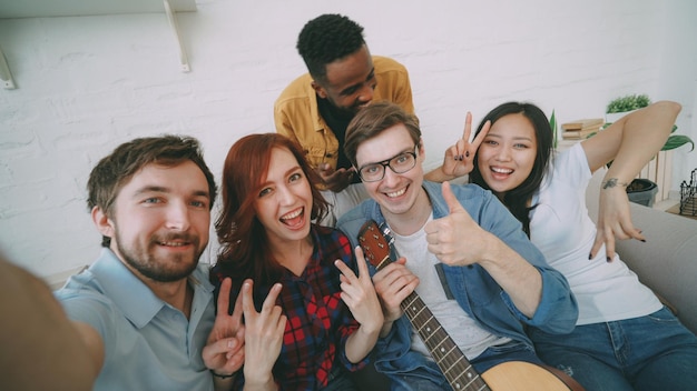 Point of view shot of multi ethnic group of cheerful friends taking selfie photos on smartphone camera while celebrating at party at home indoors