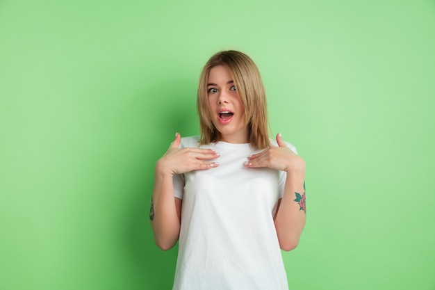 Photo pointing at herself. caucasian young woman's portrait isolated on green studio wall.