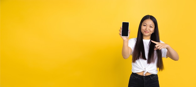 Pointing on phone. Portrait of young asian woman isolated on yellow