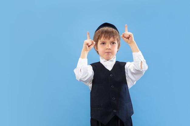 Pointing. Portrait of a young orthodox jewish boy isolated on blue studio wall.