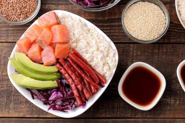 Poke bowl Hawaiian food a plate of rice salmon avocado cabbage and cheese next to sesame and soy sauce on a brown wooden table top view