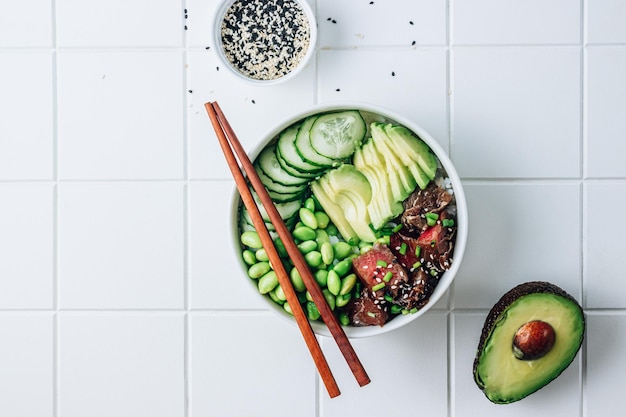 Poke bowl with raw tuna rice avocado edamame beans and cucumber in a bowl Hawaiian ahi poke bowl healthy food White ceramic tile background Top view with copy space