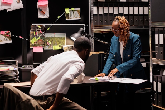 Photo police office secretary reading crime case file for detective. african american man investigator and woman assistant studying forensic expertise report and working at night time