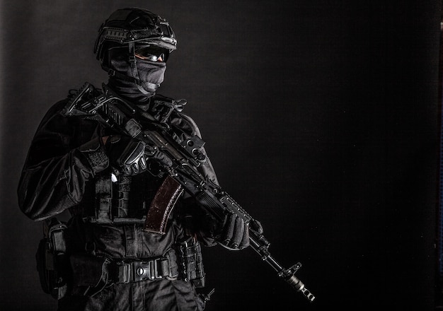 Police special forces, quick reaction team officer, tactical group fighter in black uniforms, helmet and hidden behind mask identity, armed with assault rifle, low key studio shoot on black background