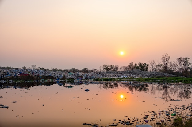 Polluted water and mountain large garbage pile and pollution at the sun is setting in the background