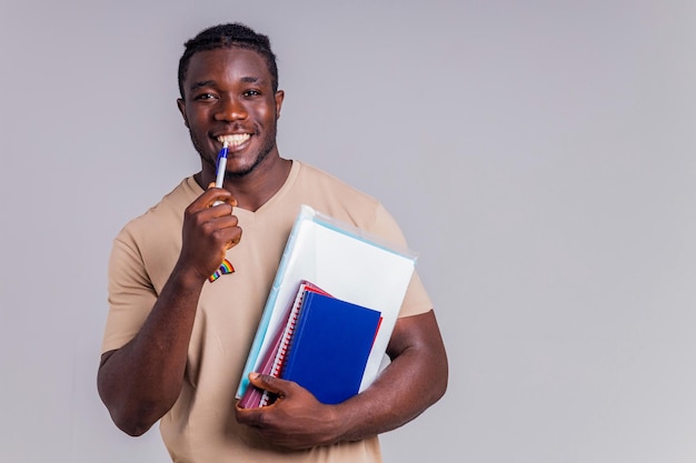 Polyethnic man in beige tshirt with an orange backpack holding book and notebooks in gray background studio