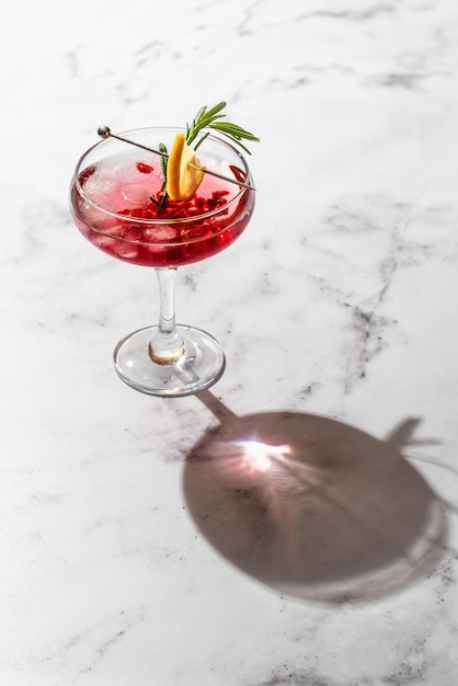 Pomegranate Christmas iced cocktail with rosemary and lemon on white marble background isolated with clear shadows in hard light space for text Xmas drink