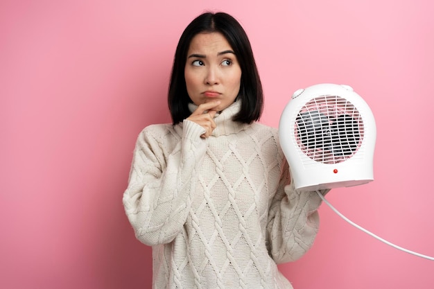 Pondering woman in warm clothes holding radiator on pink background