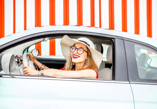 Poortrait of a young happy woman driving a car on the red wall background during the summer vacation