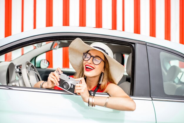 Poortrait of a young happy woman pulling out of the car window with photo camera on the red wall background during the summer vacation