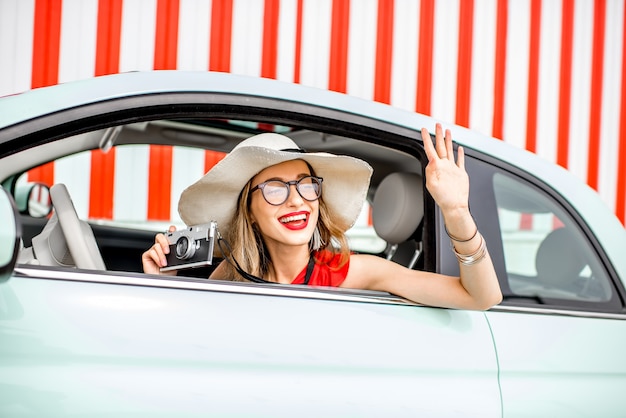 Poortrait of a young happy woman pulling out of the car window with photo camera on the red wall background during the summer vacation