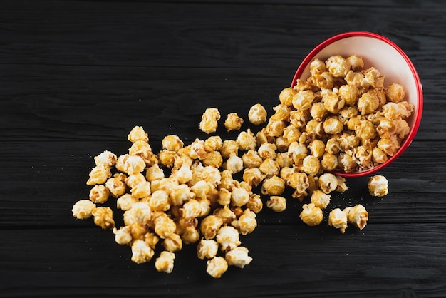 Popcorn with caramel in a red plate on a black wooden background scattered