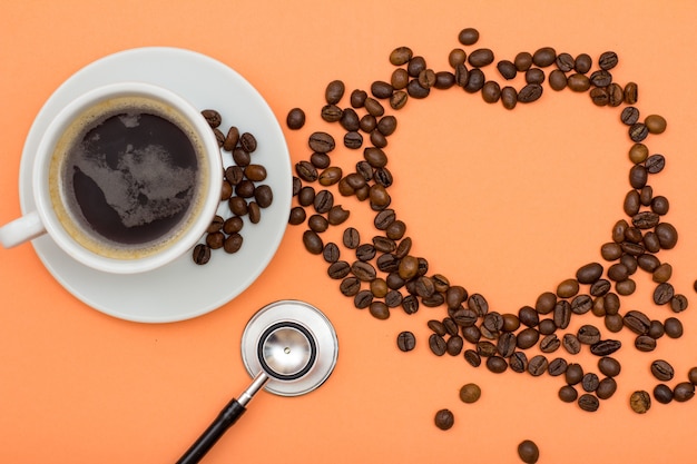 Porcelain cup of coffee on white saucer with coffee beans folded in the form of a heart and phonendoscope on peach colored background. Top view. Health care concept