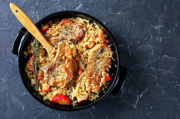 Pork Chops with Sauerkraut and Apples in a black ceramic braiser on a concrete table with a wooden spoon,