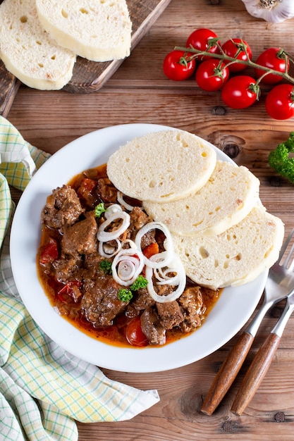 Pork goulash meat with dumplings on white plate cutlery garlic onion tablecloth in the background typical Czech food