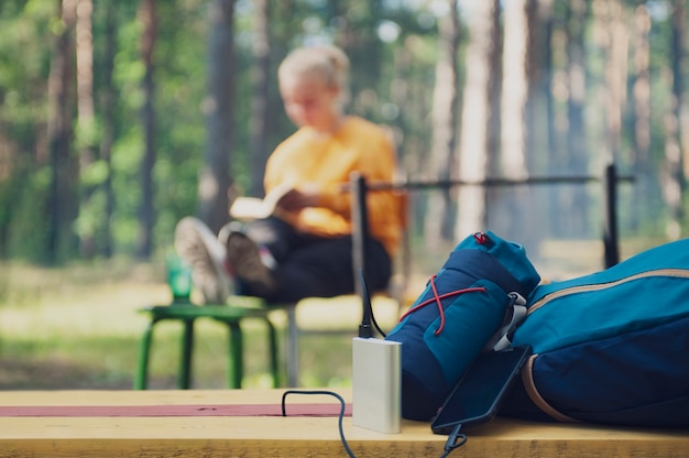 Portable Charger in Camping. Girl tourist reading a book in the woods on the background of a backpack and bank turn.