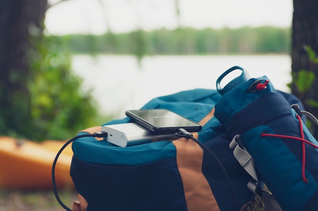 Portable travel charger. Power Bank charges Smartphone against a backdrop of journey bags, a lake and forest.