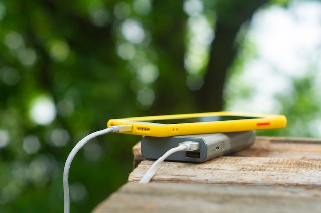 Portable travel charger. Power Bank charges a smartphone on a wooden table, in the background of nature. Concept on the theme of tourism.