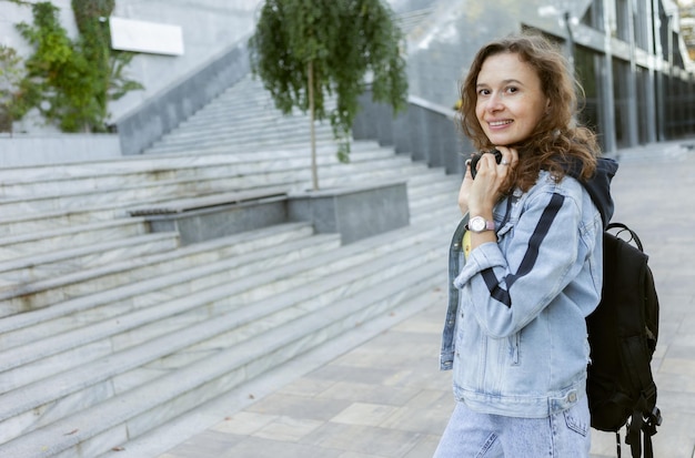 Portet charismatic curly hair woman in denim jacket with backpack and headphones outdoors in city