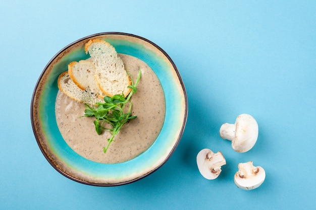 Portion of creamy mashed soup puree with champignon mushrooms, sour cream, herbs, croutons in rustic bowl on blue background