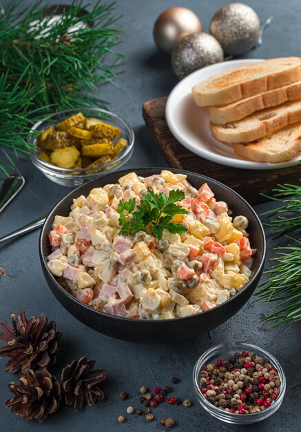 A portion of olivier salad in a black bowl against a background of pine branches and fir toys on a dark blue background. Side view, close-up.