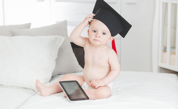 Portrait of 10 months old baby boy in graduation cap sitting with digital tablet on sofa at living room