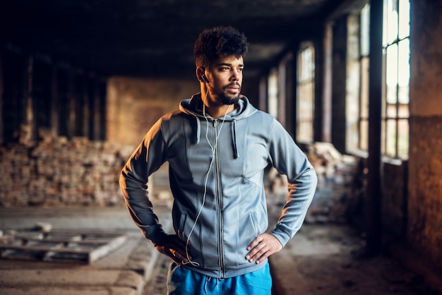 Portrait of active motivated afro-american young attractive athletic man with earphones standing inside of the abandoned place and looking far away.