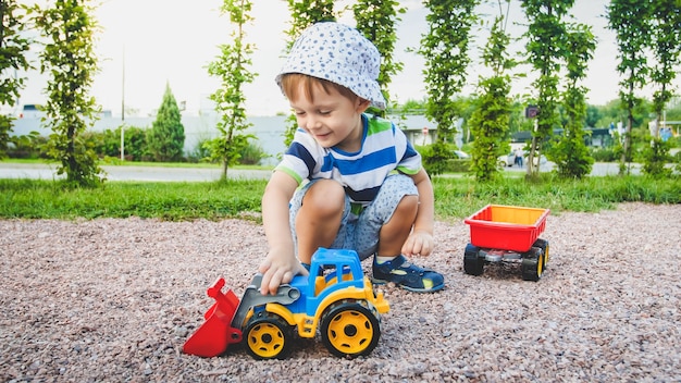 Portrait of adorable 3 years old toddler boy playing with toy truck with trailer on the playground at park