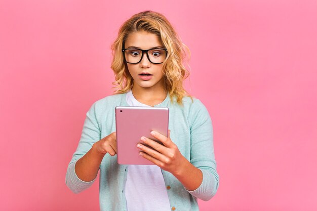 Portrait of adorable blonde kid using digital tablet device, isolated over pink background.