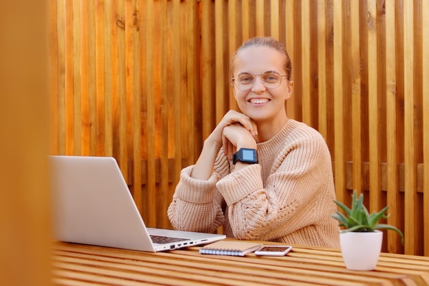 Portrait of adorable happy Caucasian woman wearing beige sweater having rest while working on laptop looking at camera with positive expression at camera posing in office against wooden wall