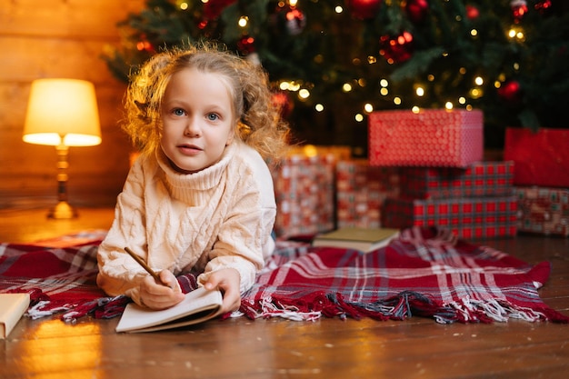 Portrait of adorable little blonde curly child girl writing letter to santa claus lying on floor on