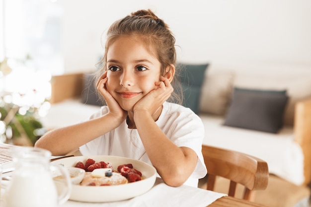 Portrait of adorable little girl sitting at table in bright kitchen while having breakfast and eating pancakes with raspberry