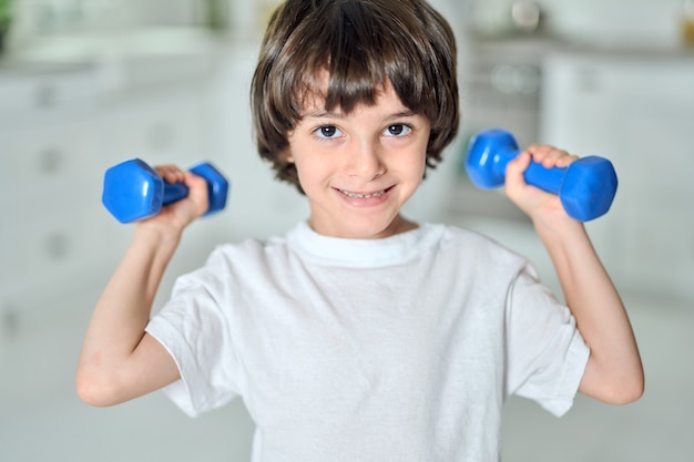 Portrait of adorable little hispanic boy smiling at camera, having workout with dumbbells in the morning at home. Childhood, healthy lifestyle concept