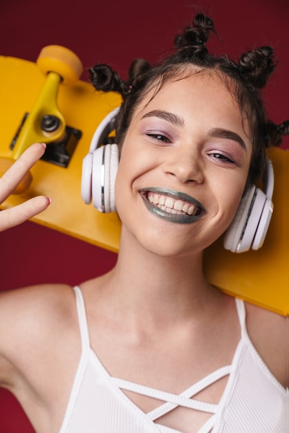 Photo portrait of adorable punk girl with bizarre hairstyle and dark lipstick holding skateboard while listening to music with headphones isolated over red wall